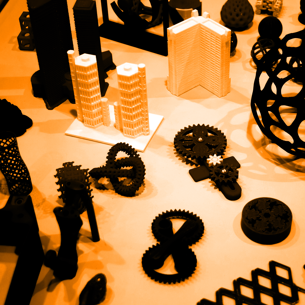 Collection of 3D-printed models including buildings, gears, and abstract shapes, displayed on a table in an orange hue.